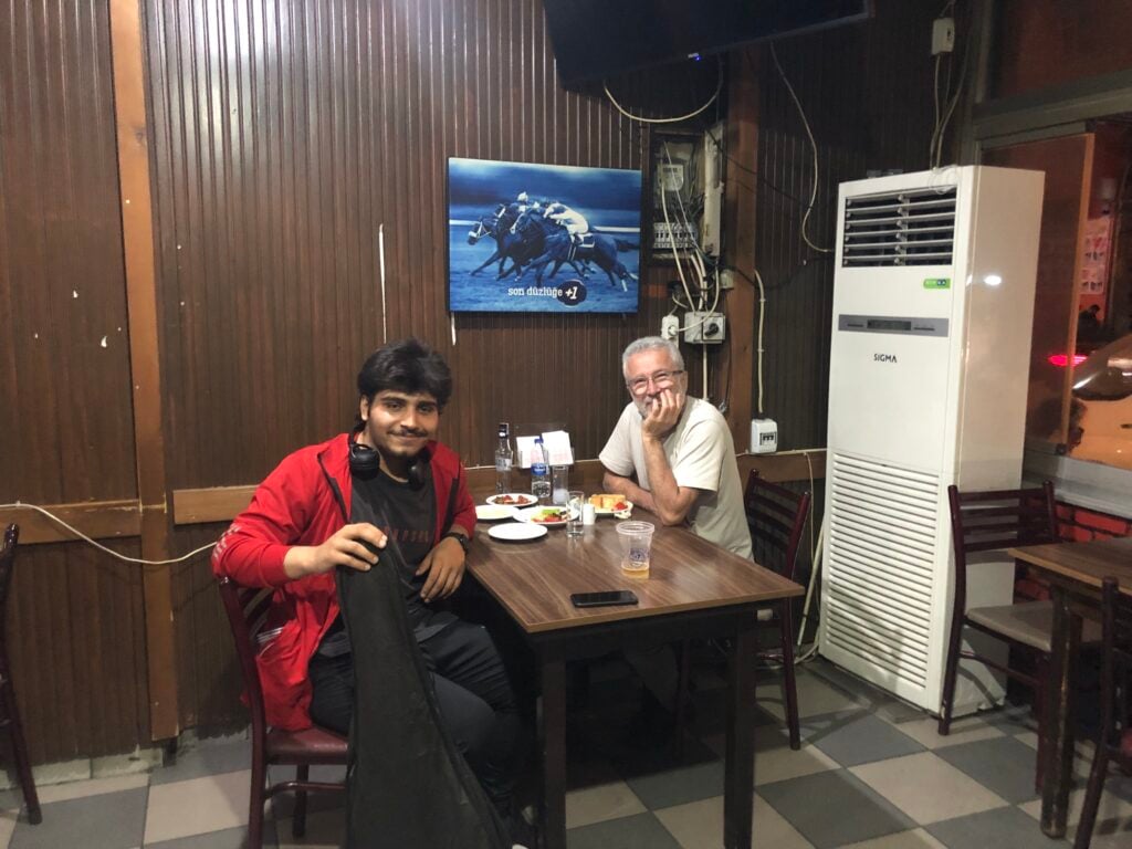 Two men sit at a dining table with plates and drinks in a wood-paneled room, a poster of galloping horses on the wall behind them, and a large white appliance to the right.