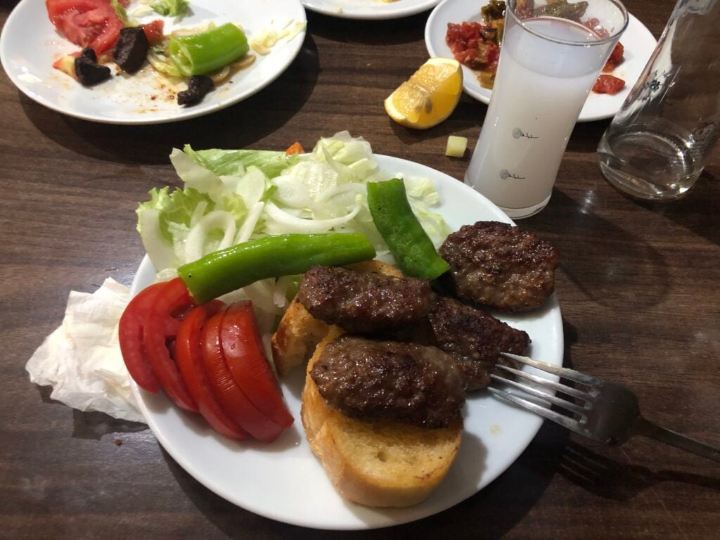 Plate with lettuce, sliced tomatoes, and a beef-topped crostini on the edge; lemon wedge and beverages nearby.