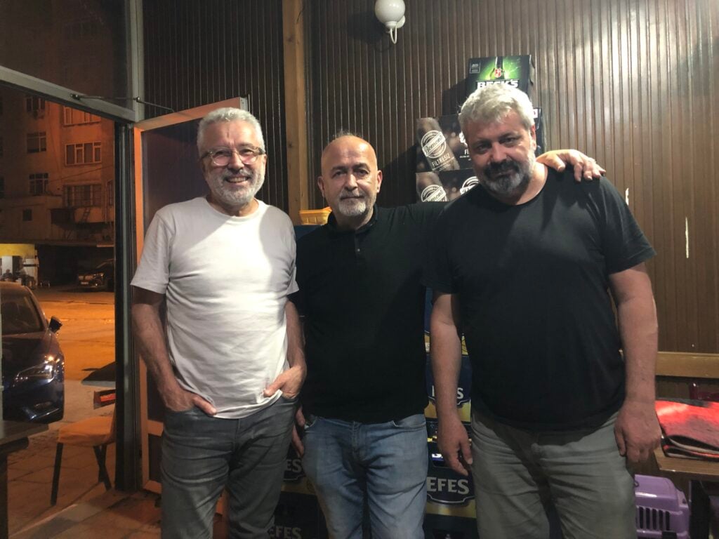 Three men stand close together and smile for a group photo indoors, wearing casual T-shirts and jeans as a nighttime street scene is visible outside.