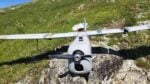 Front view of a small gray ultralight airplane with a propeller, resting on rocky ground with a grassy hillside behind.