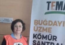 Woman in an orange safety vest stands indoors beside a large banner with Turkish protest text.