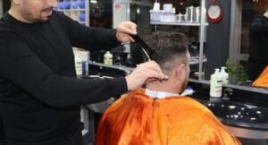 Barber trims a client's hair with scissors and a comb in a barbershop, client wearing an orange cape.