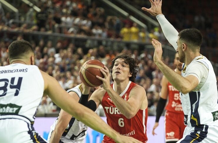 Basketball player in a red jersey with number 6 holds the ball while being defended by three white-jersey players during a crowded indoor game.