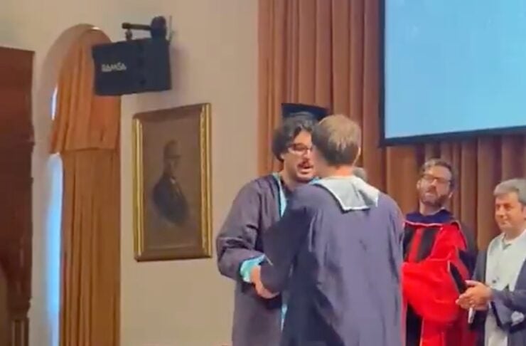 Two graduates in blue gowns embrace on stage during a ceremony, with faculty in red gown and others applauding nearby.