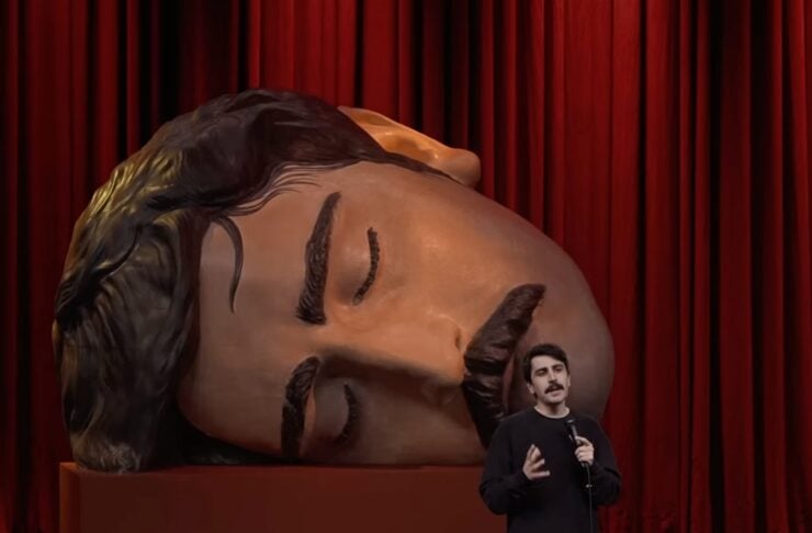 Comedian on stage with microphone beside a giant reclining sculpted head statue, red curtains in the background.