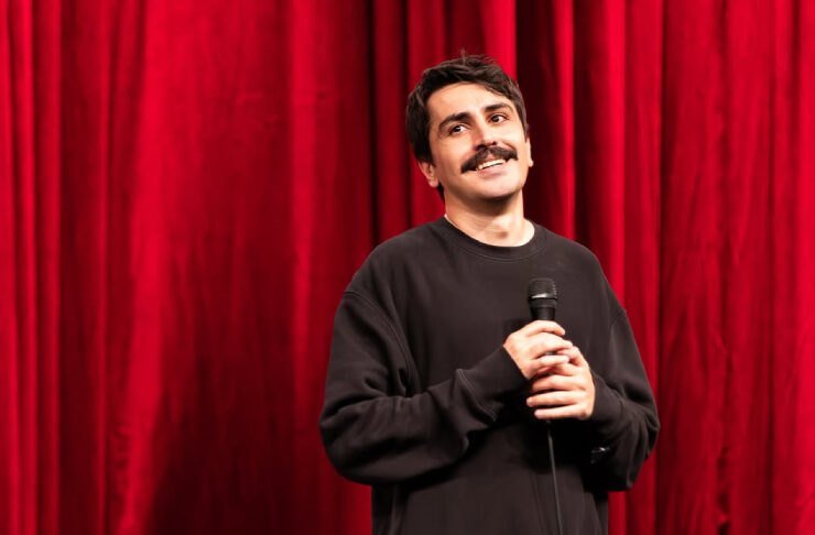 Man in black holds microphone on stage with red velvet curtain behind him.