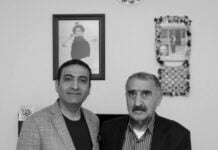 Two men in jackets posing side by side for a photo indoors, with framed pictures on the wall behind them.