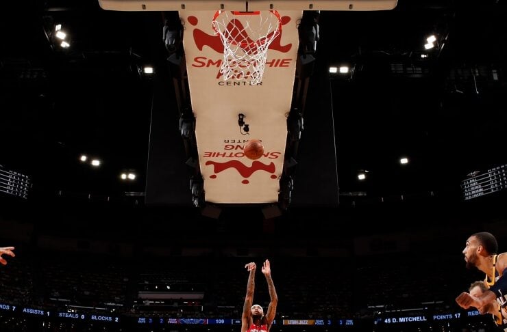 Basketball player in red jersey (#14) jumps to take a shot at the hoop while teammates and fans watch.