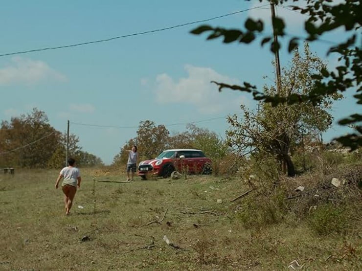 Two people in a dry, open field near a red compact car; one person walks away from the camera toward the car while another stands by the vehicle, with trees and utility wires in the background.