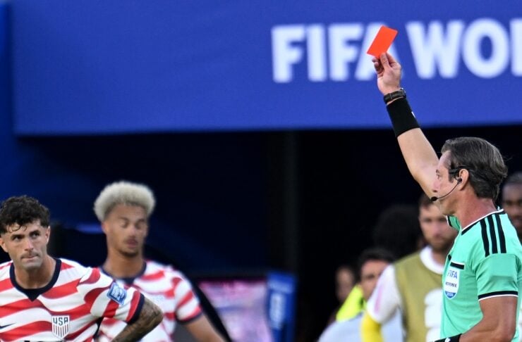 Referee in green shirt raises a red card during a USA soccer match; players in striped jerseys react in the background with 'FIFA World Cup' signage above.