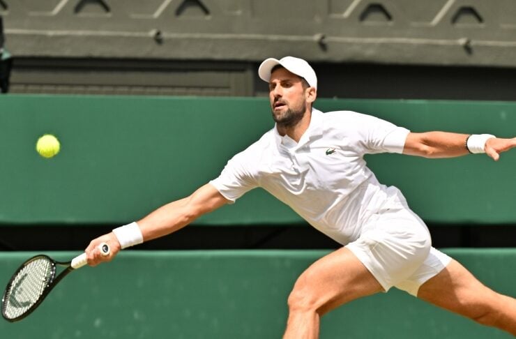 Male tennis player in a white outfit diving to hit a forehand on a grass court with a green backdrop behind him.