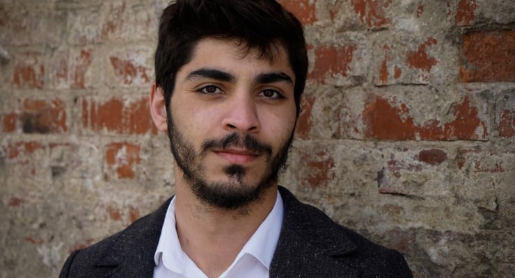 Young man with dark hair and beard, wearing a white shirt and dark blazer, stands against a weathered brick wall.