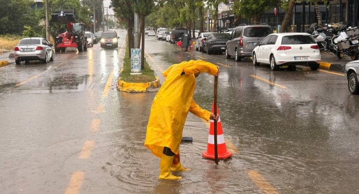 Person in a bright yellow raincoat and boots bends to adjust an orange traffic cone in a flooded city street during rain outside parked cars.