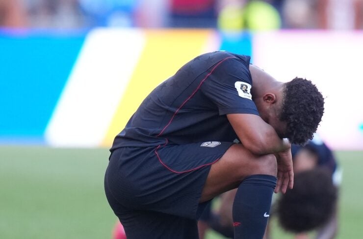 Soccer player in a dark kit kneels on the grassy field, head bowed in a moment of reflection during a game.