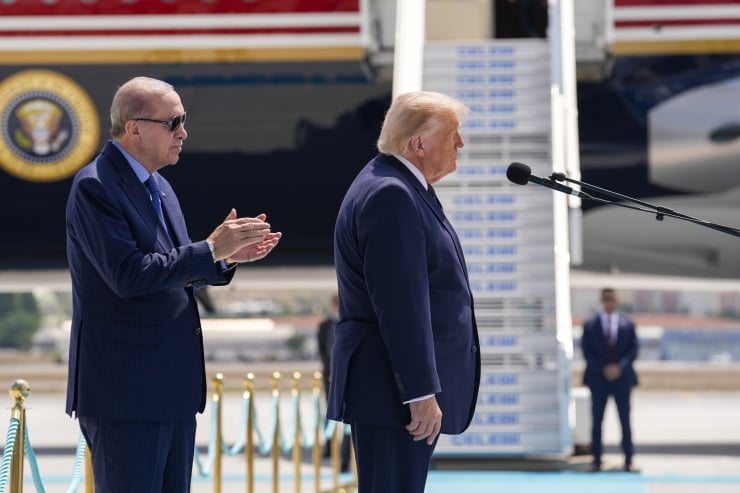 Two men in dark suits on a tarmac beside a large aircraft; one speaks into a microphone while the other claps.