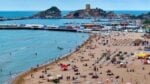 Busy beach with loungers and umbrellas, blue sea, harbor, and a rocky island with a tower in the distance.