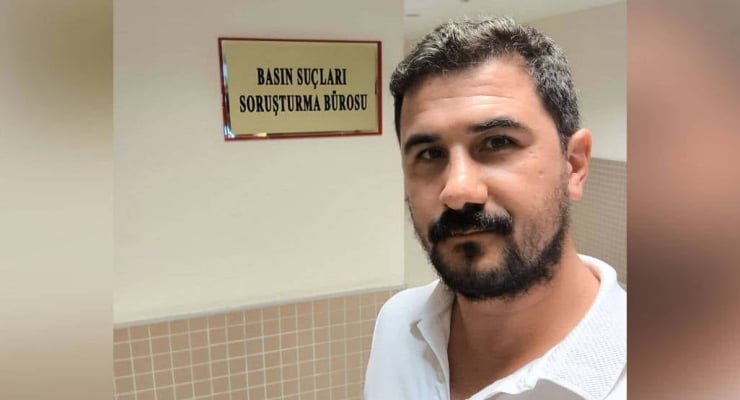 Man with short dark hair and beard in a white polo standing in a hallway beside a sign that reads 'BASIN SUÇLARI SORUŞTURMA BÜROSU'.