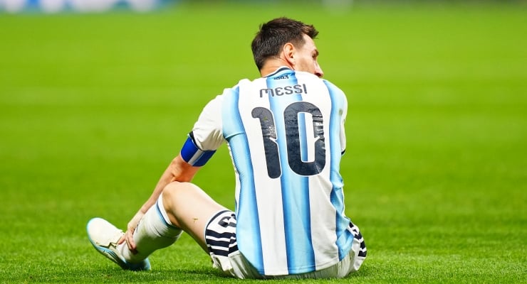 Soccer player in an Argentina jersey (number 10) sitting on a green field after a play fall-oriented moment, facing away.