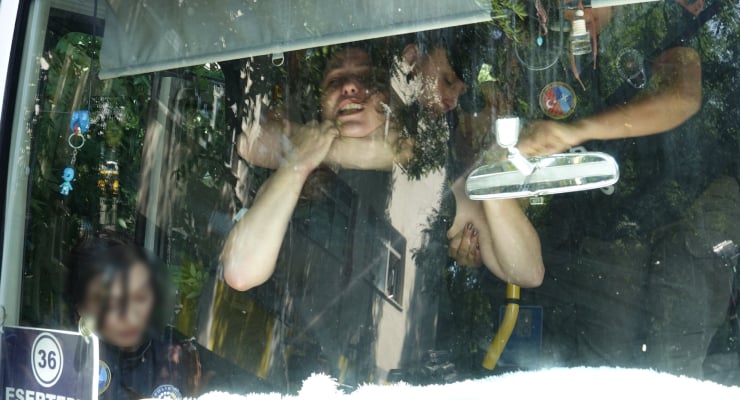 People inside a bus seen through the windshield; a man touches his neck, a blurred woman sits on the left, with trees reflected on the glass.