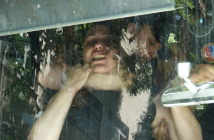 Two women in a car seen through a windshield, with reflections; one smiles while touching her neck and the other looks down.