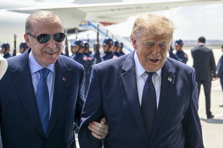 Two men in dark suits walk arm in arm on an airport tarmac, with a row of uniformed soldiers and a large aircraft in the background.
