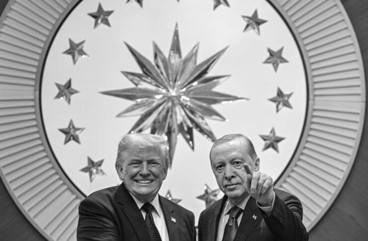 Two suited men shake hands in a formal setting with a large starburst emblem behind them; one man points toward the camera.