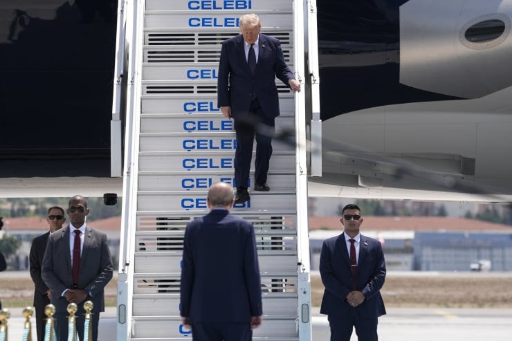 A man in a dark suit descends the stairs from a jet on a tarmac, flanked by security guards in suits.