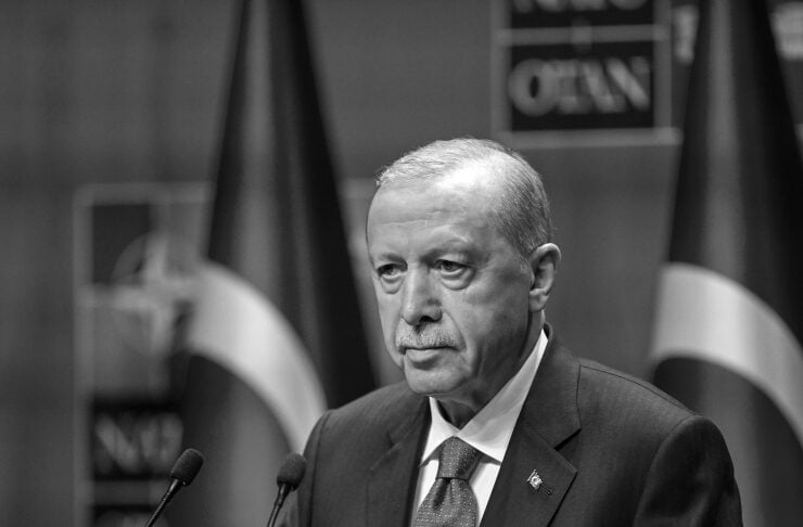 Man in a suit delivering a speech at a podium with two microphones, Turkish flags in the background.