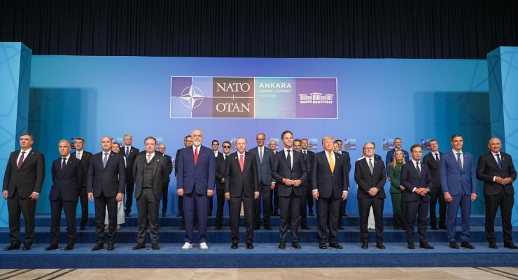 Group of world leaders in suits posing on a blue stage during the NATO summit in Ankara, with a banner displaying NATO and OTAN logos behind them.