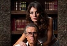 Older man in a white blazer and glasses sits at a desk while a younger woman stands behind him, hands resting on his shoulders in a library setting.