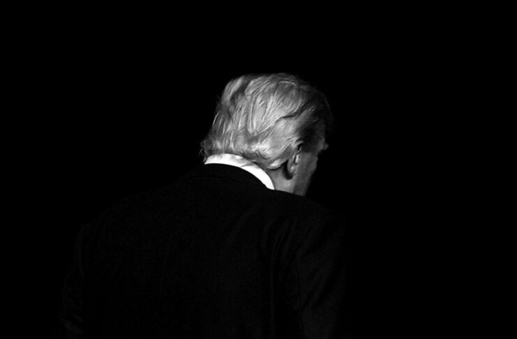 Back view of a person with light hair in a dark suit standing against a black background.