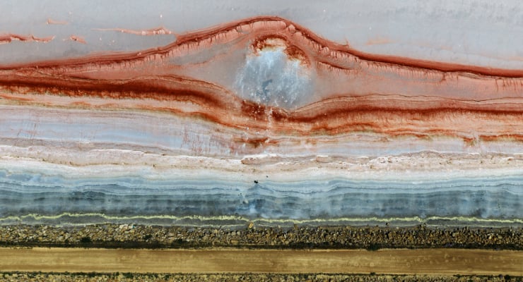 Aerial view of colorful layered mineral formations with red ridges above blue and brown bands across a flat landscape.