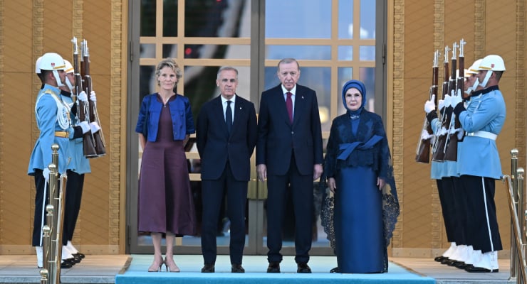 Five formally dressed figures stand on a blue carpet between two lines of uniformed guards with rifles outside a grand doorway on a ceremonial entrance.