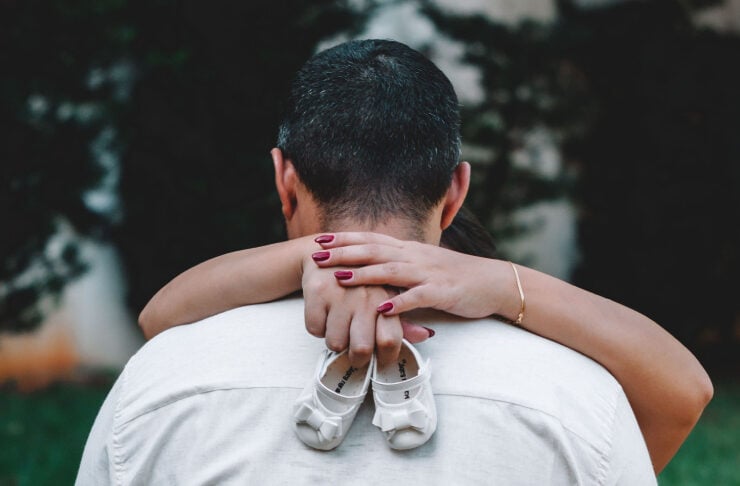 Back view of a man in a light shirt being hugged from behind by a woman's arms; white baby shoes hang from his back as a symbol of a newborn.