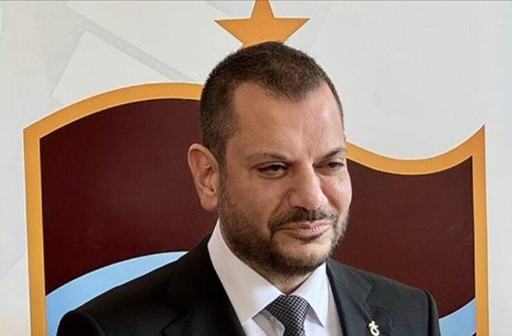 Man in a dark suit and tie stands before a maroon-and-gold shield emblem with a blue curved stripe in the background.