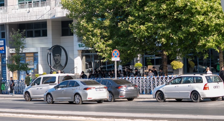 Crowd of people standing behind metal barricades along a city sidewalk in front of a white building, with several parked cars in the street and a no-parking sign nearby.