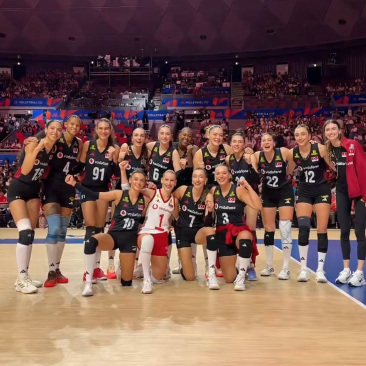 Women’s volleyball team in black uniforms huddle on the court, arms around each other, smiling after a match.