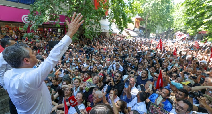 Speaker on a street stage waves to a dense outdoor crowd, many people filming with phones amid trees and banners.