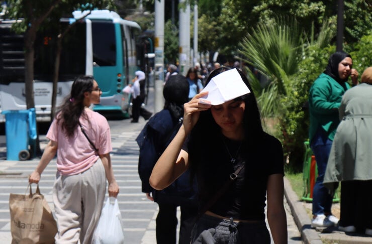 Young woman wearing a white paper hat adjusts it on her head while walking on a city sidewalk, phone in hand
