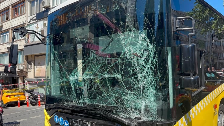 Yellow city bus with a shattered front windshield showing a web of cracks on a city street with storefronts in the background.
