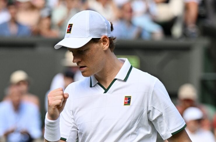 Male tennis player in a white outfit pumping his fist during a match on a grass court, crowd blurred in the background.