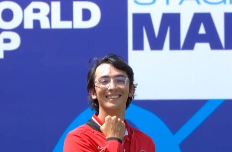 Smiling athlete in a red Turkish team jacket poses with a fist near the chin against a blue event backdrop that reads World Cup/Stage signage.