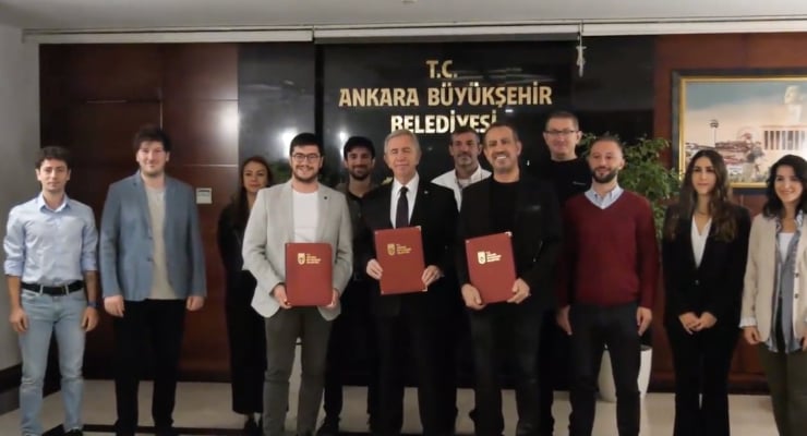 Group of professionals posing in a hallway; several men hold red folders with official logos, in front of a sign reading 'T.C. Ankara Büyükşehir Belediyesi'.