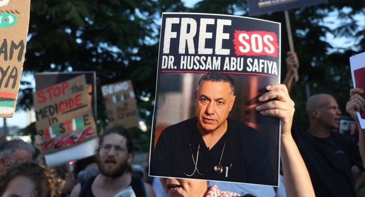 Protester holds a large sign demanding 'FREE' a doctor, with the doctor's portrait on the poster; crowd in the background.
