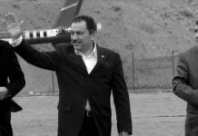 Man in a dark suit waving with his right hand outdoors, with others nearby on a dirt area.