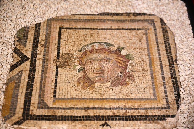 Rectangular ancient mosaic on a floor showing a central Medusa-like head surrounded by geometric borders on a pebbled surface.]?