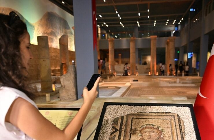 Woman in a white top and checkered skirt uses her phone to photograph a small ancient mosaic on a pedestal in a museum display area with columns in the background.