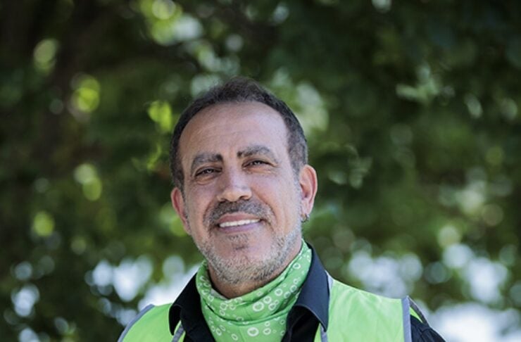 Man smiling in a neon green safety vest with a green bandana, outdoors among trees (volunteer/worker appearance).