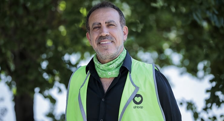 Man smiling in a lime safety vest with a green bandana around his neck, outdoors among trees.