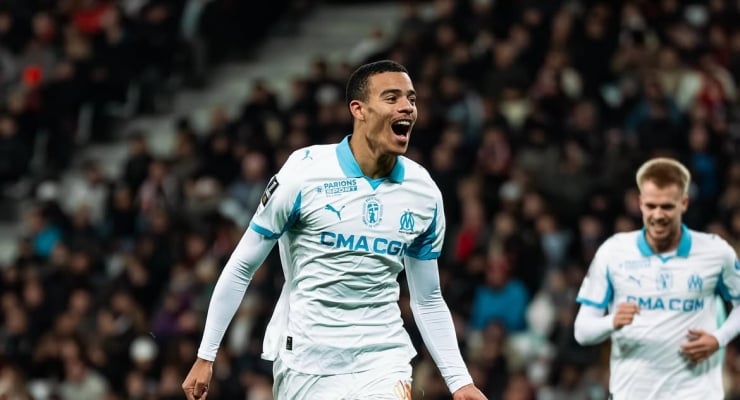 Soccer player in a white Marseille kit celebrates a goal on the field, arms outstretched with a teammate in the background.